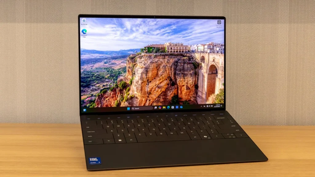 Top 10 Laptop Brands in 2026. A high-end Dell laptop opened on a light wood desk, displaying a crisp landscape wallpaper of an ancient bridge and canyon.